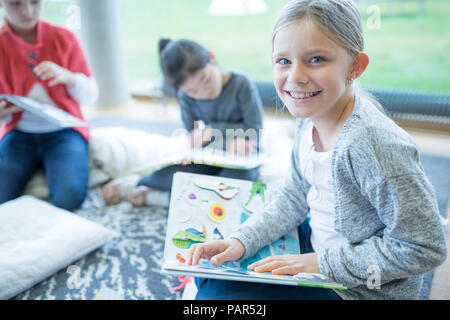 Portrait von lächelnden Schulmädchen auf dem Boden sitzend mit Buch in der Schule pause Zimmer Stockfoto