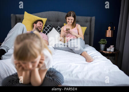 Vernachlässigte Mädchen sitzen auf dem Bett mit ihren Eltern, mit Smartphones Stockfoto