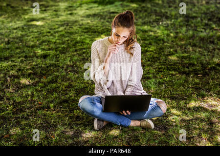 Nachdenkliche Frau sitzt auf einer Wiese mit Laptop Stockfoto