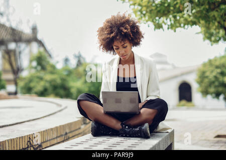 Modische junge Frau mit lockigem Haar sitzt auf der Bank mit Laptop Stockfoto