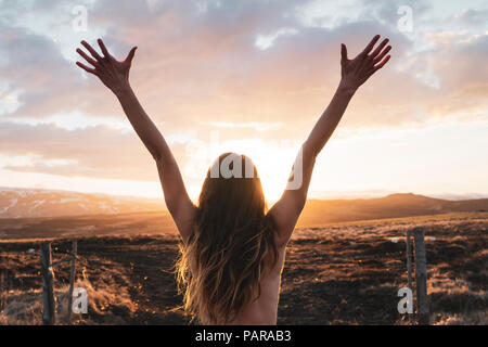 Island, junge Frau mit erhobenen Armen bei Sonnenuntergang Stockfoto