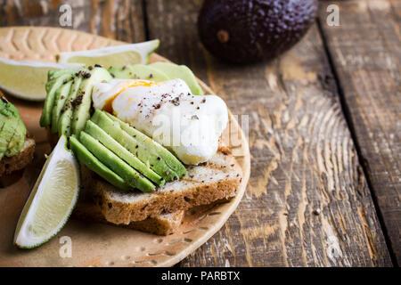 Pochierte Eier in Scheiben geschnitten und Avocado auf Vollkornbrot Toast serviert mit Kalk auf Keramikplatte auf rustikalen Holztisch, vegetarische Sandwiches Stockfoto