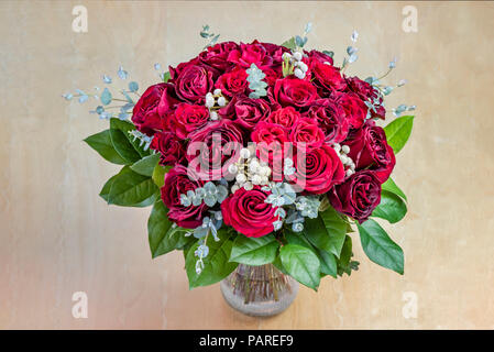 Ansicht von oben auf ein Strauß roter Rosen mit grünen Blättern in einem Glas Vase auf einem Holztisch Stockfoto