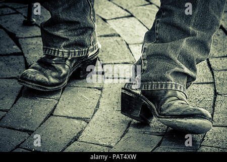 Eine Nahaufnahme der Füße eines Mannes in abgetragene Cowboystiefel mit großen Fersen und Jeans auf gebogener Ziegelstein weg in Schwarz und Weiß Stockfoto