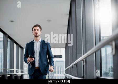Junge Unternehmer in einer Passage mit Handy, Ohrhörern und rollenden Koffer unterwegs. Stockfoto