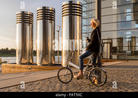 Ältere Frau mit Takeaway Kaffee und City Bike mit einer Unterbrechung bei Sonnenuntergang Stockfoto