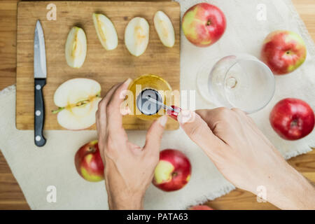 Öffnen der Flasche Apfel cidre trinken, Ansicht von oben. Sicht der Hand mit Dosenöffner, Vorbereitung ein Getränk von Apfelwein auf rustikalen Holztisch mit reifen Appl Stockfoto