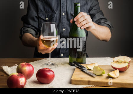 Männliche Hände, die Flasche premium Cider auf rustikalen Holztisch. Schöne Eis kalte Flasche Apfelwein, mit reife Äpfel im Hintergrund Stockfoto