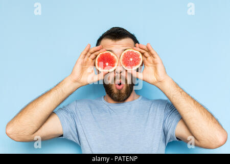 Junge bärtige Mann mit Schichten von Grapefruit vor seinen Augen. Hellblauer Hintergrund, überraschten Ausdruck auf dem Gesicht des Menschen.. Stockfoto