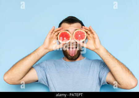 Junge bärtige Mann mit Schichten von Grapefruit vor seinen Augen. Hellblauer Hintergrund, der Mensch, seine Zunge. Stockfoto