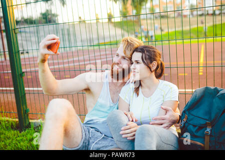 Ein mans Paar rote Haare und Bart und Frau Erholung nach Sport im Freien auf Gras im Hintergrund des Stadions. Der Kerl hält red Smartphone auf outstre Stockfoto