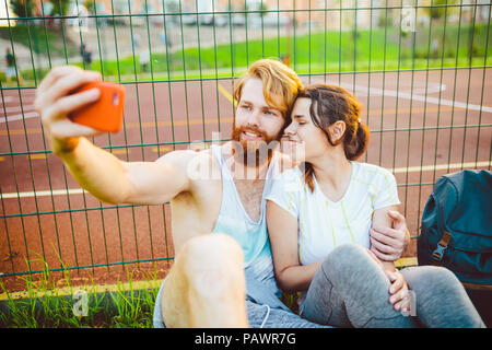 Ein mans Paar rote Haare und Bart und Frau Erholung nach Sport im Freien auf Gras im Hintergrund des Stadions. Der Kerl hält red Smartphone auf outstre Stockfoto