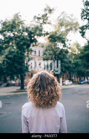 Junge Frau mit dicken lockigen Haar an der Straße und um die Stadt zu Fuß zu erkunden. Ansicht der Rückseite des unkenntlich Weibliche trägt blaue Shirt stehend Stockfoto