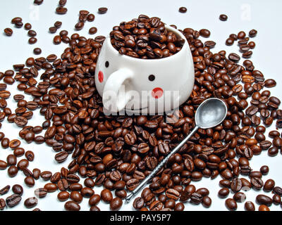Eine Kaffeetasse mit Körnern aus gerösteten Kaffee und einem Löffel daneben sind auf gerösteten Kaffeebohnen platziert Stockfoto