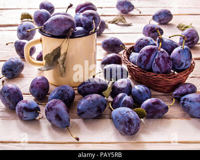 Blau große Pflaumen saftige süße Frucht für diätetische Ernährung liegen in einem gelben Metall Becher und in einem Weidenkorb auf eine Planke Hintergrund Stockfoto