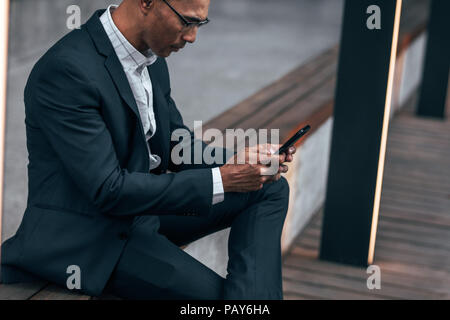 Unternehmer mit der Texteingabe im Handy sitzen auf Seite Geländer im Freien. Unternehmer Sitzen im Freien verwalten Arbeiten mit Handy. Stockfoto