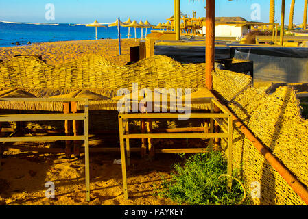 Eine Reihe von Stroh Sonnenschirme gegen Überhitzung und Sonnenliegen am Strand vor einem blauen Himmel und das blaue Meer zu schützen. Das Konzept der Sommerferien, Stockfoto