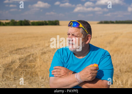 Outdoor Portrait eines bärtigen Älterer in der Sonnenbrille, die Aprikosen in der Hand gegen den blauen bewölkten Himmel und landwirtschaftlichen Bereich Stockfoto