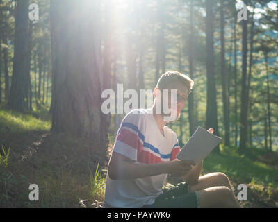 Junge handscome Mann in zwanglosen Sitzen auf dem Boden im Wald und Poesie im Notebook an einem sonnigen Tag Stockfoto