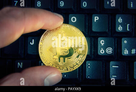 Mit zwei Fingern halten, einen goldenen Bitcoin über die Tastatur eines Computers. Stockfoto