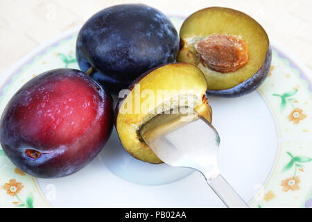 Ganze und eine halbierte Reif natürliche Pflaumen, saftig und frisches Obst auf dem Tisch mit Löffel in der Mitte Stockfoto