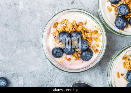 Frühstück aus frischen Joghurt mit hausgemachtem Müsli und Heidelbeeren in Gläsern auf grauem Hintergrund Stockfoto