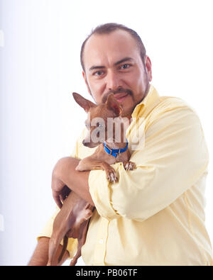 Studio Portrait auf einen weißen Hintergrund von einem Mann in einem gelben Hemd, mit einem Chihuahua Hund in seine Arme Stockfoto