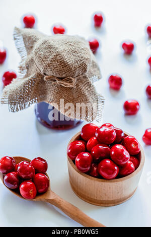 Rote reife Kirschen mit Jam jar und Holzlöffel auf weißem Hintergrund. Flach. Essen Konzept. Stockfoto