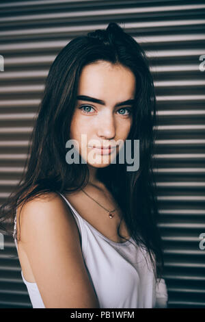 Portrait von cute girl mit langen dunklen Haaren und blauen Augen. Junge Frau tragen weiße oben stehend über braunen Hintergrund an Kamera und Lächeln suchen. Stockfoto