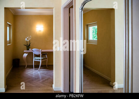 Ein Vorraum mit einem kleinen Schreibtisch und einer Orchidee gehört zu einem Zimmer im Hotel Lenkerhof in Lenk, Schweiz. Spiegeltüren und Fischgratparkettböden prägen den Flur. Stockfoto