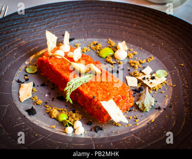 Rindertatar mit Armagnac, ein Gericht im Restaurant Spettacolo des Hotels Lenkerhof in Lenk, Schweiz. Dunkler Brotkrümel, cremefarbene Perlen und dünne Chips umgeben das Fleisch auf einem schiefergrauen Teller. Stockfoto