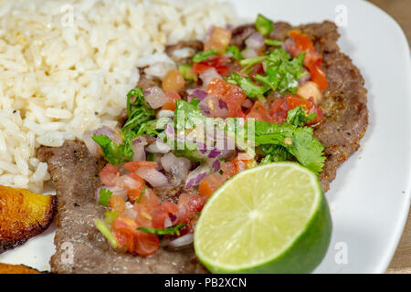 Ein Steak in Pico de Gallo von Kochbananen und weißen Reis auf einem weißen Teller umgeben. Kubanisches Essen. Stockfoto