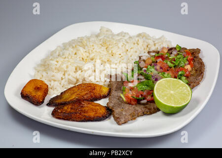 Ein Steak in Pico de Gallo von Kochbananen und weißen Reis auf einem weißen Teller umgeben. Kubanisches Essen. Stockfoto