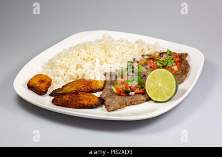 Ein Steak in Pico de Gallo von Kochbananen und weißen Reis auf einem weißen Teller umgeben. Kubanisches Essen. Stockfoto