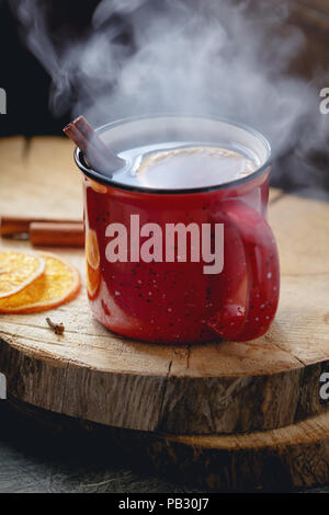 Glühwein in einem roten Keramik Becher über rustikalen hölzernen Brettern umgeben Gewürze. Stockfoto