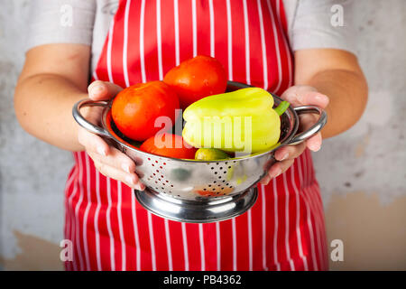 Frau in einem roten Schürze mit einem Sieb mit Gemüse für Salat. Das Kochen. Stockfoto