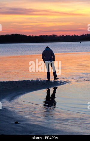 Silhouette einer Person, die am Rande des Wassers von einem Strand bei Sonnenuntergang einen Fuß anheben mit einem nassen Schuh aus dem Wasser Stockfoto