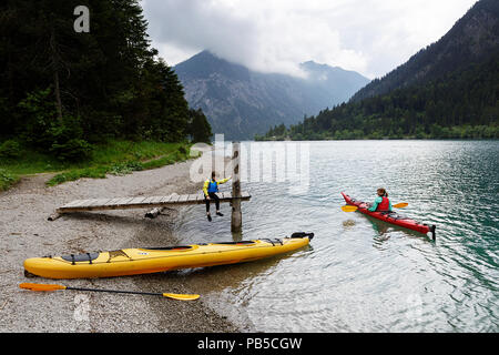 Mutter in einem Kajak im Gespräch mit ihrem Sohn sitzen auf dem hölzernen Pier mit einem Kajak am Ufer am Plansee, Österreich geparkt Stockfoto