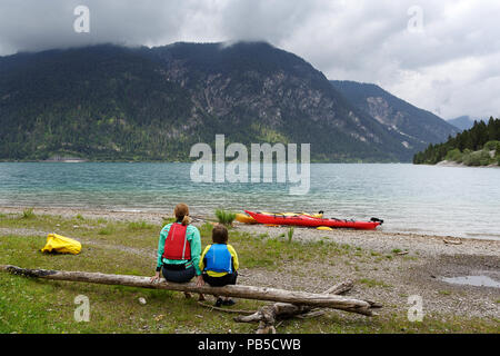 Mutter und Sohn mit allen Kajak Ausrüstung auf einem getrockneten Log nach dem Kajakfahren auf am Plansee, Österreich sitzen Stockfoto