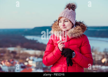 Frau Anwendung Lippenbalsam beim Spaziergang in einem Wintertag. Das Tragen der roten Mantel und Mütze. Stadt im Hintergrund Stockfoto