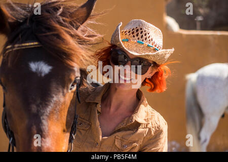 Fröhliche junge Dame in Landschaft bereiten ihre schönen braunen Pferd zu reiten. weißes Pferd im Hintergrund. Sonnenlicht Farben und Lebensstil im Freien wit Stockfoto