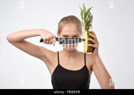 Portrait der blonden Frau, die Ananas und Messer in Händen hält gegen den weißen Hintergrund Stockfoto