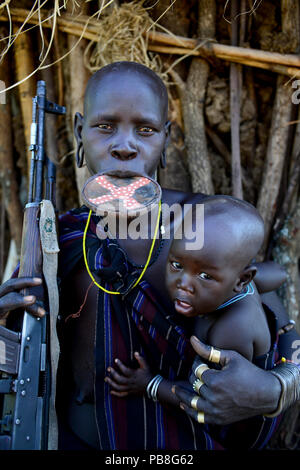 Mursi Frau mit Lehm Lippe Platte, mit Gewehr und Sohn. Mago National Park. Omo Valley, Äthiopien. Stockfoto