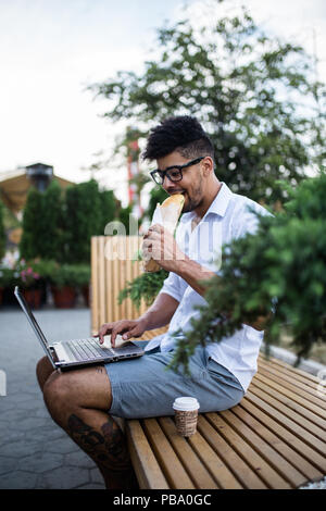 Junge hübsche Afro-amerikanische Mann mit Brille sitzen auf der Straße mit Laptop und essen Sandwich. Businesss Konzept. Stockfoto