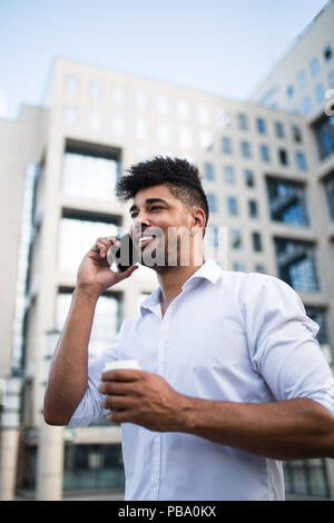 Junge hübsche Afro-amerikanische Mann stand vor der riesigen, modernen Geschäftsgebäude, Lächeln, Kaffee und Gespräch am Handy. Stockfoto