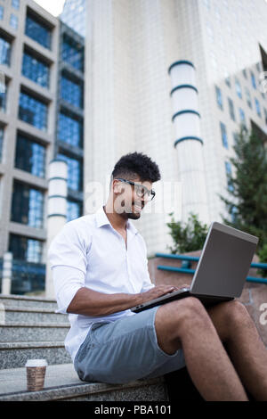 Junge hübsche Afro-amerikanische Mann vor der riesigen, modernen Firmengebäude sitzen und dabei einige Arbeiten am Laptop. Stockfoto