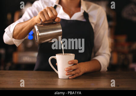 Frau mit Schürze gießen Milch in eine Tasse Kaffee, Espresso. Professionelle barista Kaffee im Cafe. Stockfoto