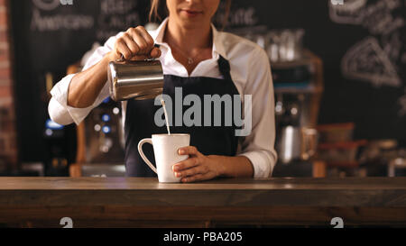 7/8 shot der weiblichen Barista eine Tasse Kaffee steht hinter cafe Zähler. Junge Frau gießen Milch in eine Tasse Kaffee. Stockfoto