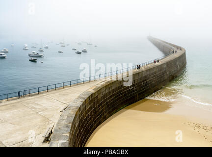 Menschen Flanieren auf der Mole in der ummauerten Stadt Saint-Malo in der Bretagne, Frankreich, ein sonniger und nebligen Tag mit Boote im Hafen Stockfoto