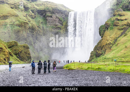 Skogafoss, Island - 14. Juni 2018: Wasserfall, Felswand, grünes Gras Hügel, viele Touristen, Menschen zu Fuß auf Schotterpiste, Nebel, Wasser fallen aus Klippe Ich Stockfoto